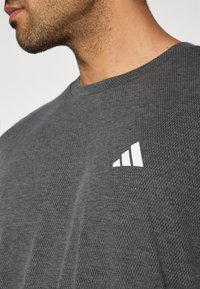 Γκρι μπλουζάκι από διαπνέον δίχτυ με στρογγυλή λαιμόκοψη, που διαθέτει λευκό λογότυπο με τρεις ρίγες στο αριστερό στήθο�ς. Η υφή είναι ελαφριά και επιτρέπει την αναπνοή.