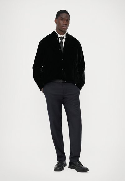 Black velvet jacket with a collar, front buttons, and a pocket. Paired with dark trousers and black lace-up shoes. Clean design and textures.