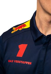 Niebieska koszulka polo z dużą czerwoną cyfrą "1" oraz napisem "MAX VERSTAPPEN" wydrukowanym na czerwono. Żółte logo Red Bull na ramieniu.