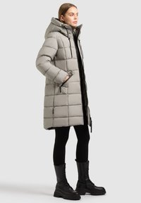 Vadderad grå jacka med huva, kviltad design och sidofickor, tillsammans med svarta leggings och snörda svarta stövlar.