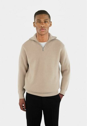 Beige ribbad tröja med halv dragkedja och avslappnad passform, med långa ärmar och en texturerad tyg. Bärs med mörka byxor.
