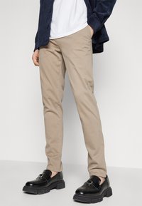 Beige tapered chino pants with a smooth texture. Paired with black chunky loafers featuring buckle detail. White t-shirt under a navy shirt.