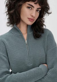 Pull en tricot vert clair avec col montant et fermeture éclair à moitié. Le tissu texturé présente des détails côtelés sur les manches pour une dimension supplémentaire.