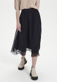 Black, flowing midi skirt with a textured waistband. Sheer fabric creates a layered effect. Worn with cream ankle boots.