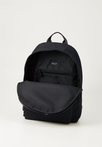 Mochila de tela negra con cremallera superior, solapa frontal, bolsillo interior y etiqueta "REPLAY" en el interior. Textura suave en toda la superficie.