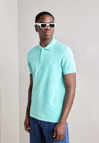 Polo bleu clair avec un col, à manches courtes et un logo brodé. Associé à un jean bleu et complété par des lunettes de soleil blanches.