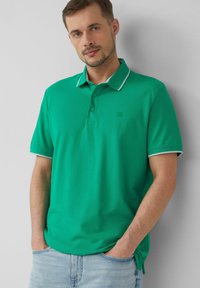 Zielona koszulka polo z prążkowanym kołnierzykiem z białymi akcentami, krótkimi rękawami i subtelnym logo na klatce piersiowej. Materiał wydaje się gładki.