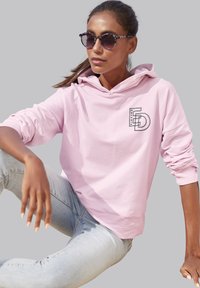 Felpa rosa chiaro con grafica frontale, caratterizzata da un cappuccio con cordoncino e polsini a coste, abbinata a jeans blu chiaro e occhiali da sole con motivo.