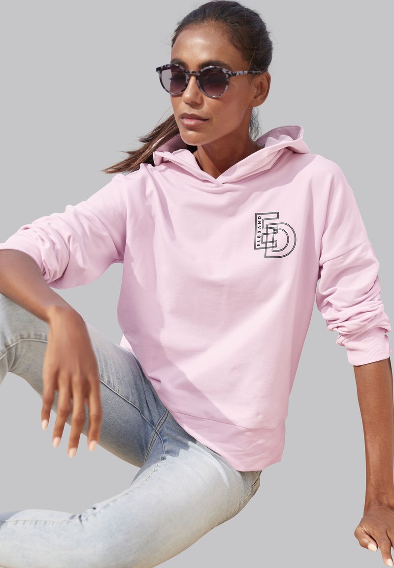 Felpa rosa chiaro con grafica frontale, caratterizzata da un cappuccio con cordoncino e polsini a coste, abbinata a jeans blu chiaro e occhiali da sole con motivo.