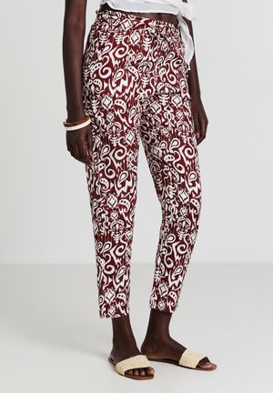 Donna che indossa pantaloni a fantasia bordeaux e bianchi, sandali beige e una maglietta cropped bianca annodata, in piedi su uno sfondo semplice.
