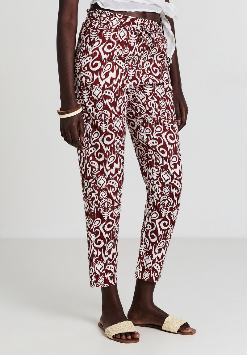 Donna che indossa pantaloni a fantasia bordeaux e bianchi, sandali beige e una maglietta cropped bianca annodata, in piedi su uno sfondo semplice.