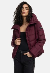 Bordeaux pufferjack met een capuchon, voorzien van een ritssluiting, een doorgestikt ontwerp en elastische manchetten. Zwarte broek maakt de outfit compleet.