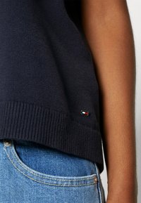 Granatowy sweter dzianinowy z prążkowanym dołem i małą metką logo w czerwonym, białym i niebieskim kolorze. W połączeniu z jasnoniebieskimi dżinsami.