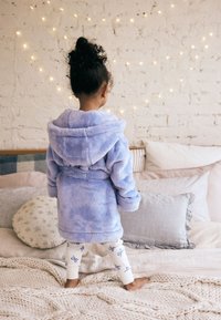 Peignoir de bain bleu et duveteux avec une capuche. Pantalon blanc orné de motifs de nœuds bleus. Enfant debout sur un lit parmi des coussins décoratifs.