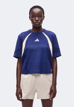 Γυναίκα με μπλε αθλητικό μπλουζάκι Adidas από δίχτυ και ανοιχτό μπεζ σορτς, που στέκεται μπροστά σε ένα απλό ανοιχτό φόντο.