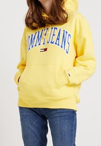 Felpa gialla con stampa frontale "TOMMY JEANS" in blu. Caratterizzata da una tasca e polsini a coste, realizzata in tessuto morbido. Abbinata a jeans blu.