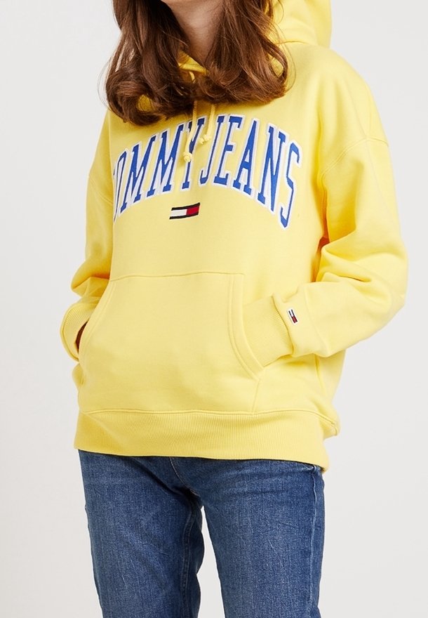 Felpa gialla con stampa frontale "TOMMY JEANS" in blu. Caratterizzata da una tasca e polsini a coste, realizzata in tessuto morbido. Abbinata a jeans blu.
