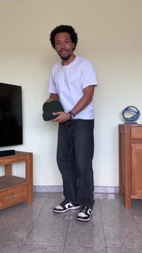 Πράσινο πλεκτό σκουφάκι στα χέρια, ραβδωτή υφή. Άτομο με λευκό T-shirt και σκούρα τζιν στέκεται δίπλα σε μια τηλεόραση και ξύλινα έπιπλα. Μαύρα αθλητικά παπούτσια.