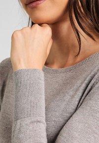 Harmaa sydämen muotoisella kaula-aukolla varustettu villapaita, jossa on joustavat hihansuut. Pehmeä rakenne näkyy, ja kangas on sileä käsivarren ja olkapään alueella.