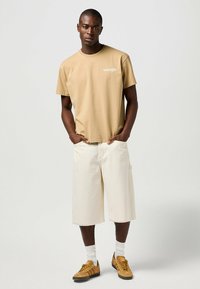Camiseta corta de color beige claro con el logo de "Wrangler", combinada con pantalones cortos color crema hasta la rodilla. El calzado consiste en zapatillas marrones y calcetines blancos.