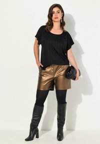Zwarte top met korte mouwen en metallic versieringen, gecombineerd met glanzende bronzen shorts, zwarte opaque panty’s en zwarte laarzen tot de knie.