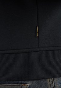 Marineblauwe sweatshirt met een geribde onderrand en manchetten, voorzien van een zwart label met gele tekst. Katoenmix, zachte textuur, minimalistisch ontwerp.