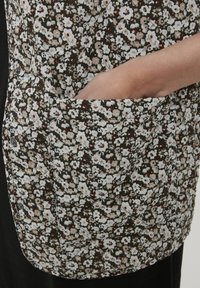 Ręka sięga do dużej kieszeni czarno-biało-brązowego ubrania z kwiatowym haftem i zapięciem na zatrzaski.