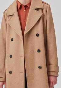 Cappotto cammello doppiopetto con bottoni neri, ampie rever, e una texture morbida. L'interno presenta una camicia arancione sotto.