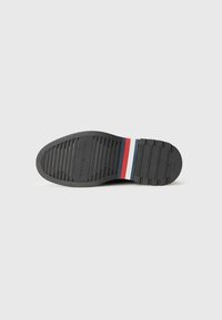 Suela de zapato de goma negra con agarre texturizado, con franjas blancas, azules y rojas en el talón. Superficie lisa y diseño curvado.