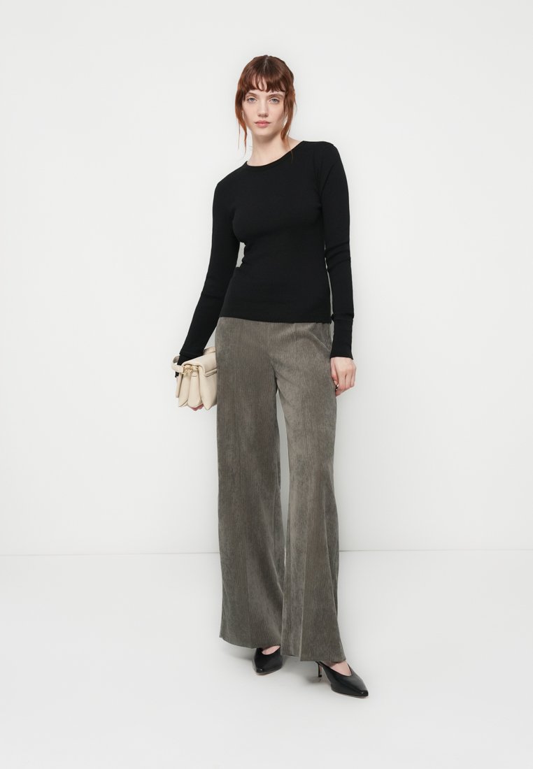 Haut noir à manches longues, ajusté. Pantalon large en velours côtelé vert olive. Le modèle tient un sac à main beige clair et porte des chaussures à talons hauts noires.