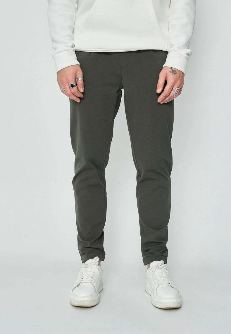 Homme portant un pantalon slim noir, des baskets blanches, un sweat-shirt blanc et plusieurs bagues aux deux mains, se tenant devant un fond blanc.
