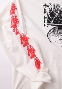 Λευκό μακρυμάνικο μπλουζάκι με κόκκινα γραφικά ενός σχήματος με τη λέξη «FORMER» στο αριστερό μανίκι και μαύρο αφηρημένο σχέδιο στο μπροστινό μέρος.