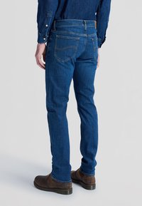 Homme portant un jean en denim bleu et une chemise en denim bleu assortie, vu de dos, associé à des bottines en cuir marron.