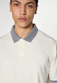 Μπλουζάκι πόλο σε κρεμ απόχρωση με υφασμάτινο γκρι γιακά, δύο κουμπιά, λεπτό λογότυπο στο στήθος και κοντά μανίκια. Ομαλή υφή υφάσματος.