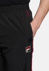 Zwarte sportbroek met een elastische tailleband, voorzien van rode zijstrepen en een FILA-logo op de linker bovenbeen.