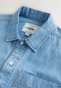 Chemise en denim bleu clair avec col boutonné, poche avant et boutons blancs. Présente des surpiqûres jaunes en guise d'accent.