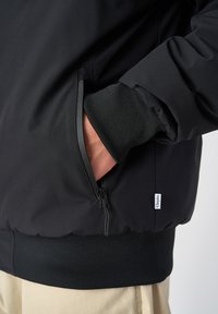 Schwarze Jacke mit einer Reißverschlusstasche, gerippten Bündchen und Saum. Glattes Material und minimalistisches Design mit einem kleinen Logo-Tag an der Seite.