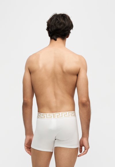 Homme debout, de dos, portant un boxer blanc avec une ceinture à motif doré sur un fond uni.