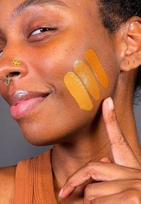 Tre fargeprøver av foundation i varme ambertoner påført kinnet til en person, med en finger som peker mot dem. Huden har en jevn tekstur.