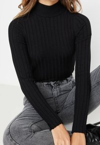Schwarzer gerippter Rollkragenpullover mit langen Ärmeln. Weiches Material, figurbetonter Schnitt und vertikale Streifen erzeugen eine strukturierte Silhouette. Kombiniert mit grauen Jeans.