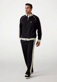 Trening negru cu accente crem; jacheta cu fermoar are un guler înalt. Pantalonii au dungi laterale. Purtați cu adidași negri.