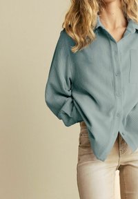 Lyseblå, struktureret skjorte med knapper, lange ærmer og en lomme, kombineret med lyse beige, tætsiddende bukser.