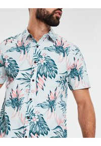 Chemise à manches courtes avec un fond bleu clair et un motif coloré de feuilles tropicales et de fleurs. Des boutons marron ajoutent du détail.