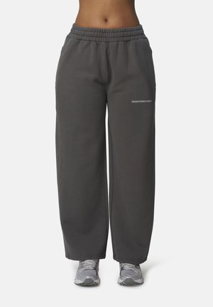 Personne portant un pantalon de survêtement gris foncé à jambes larges avec une taille élastique et des chaussures de course grises, visible de la taille jusqu’aux pieds.