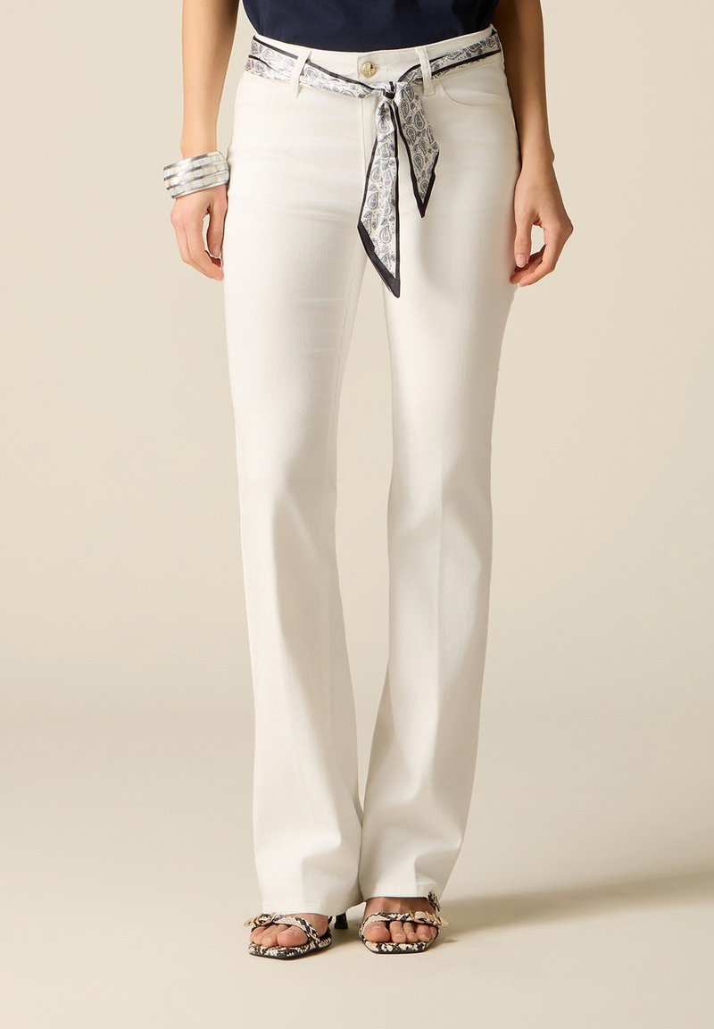 Osoba ubrana w białe spodnie dzwony z paskiem chusty we wzór oraz pasujące sandały na obcasie, stojąca na neutralnym tle.