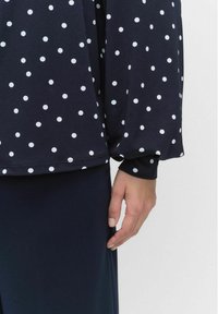 Navyblauwe blouse met witte stippen, zachte stof, lange mouwen en een ontspannen pasvorm, gecombineerd met een donkere rok, waarbij een deel van een hand zichtbaar is.