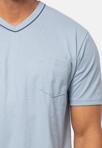 Ljusblå bomulls-t-shirt med V-ringning, korta ärmar och en bröstficka på vänster sida. Har en kontrasterande mörkblå kant längs ringningen.