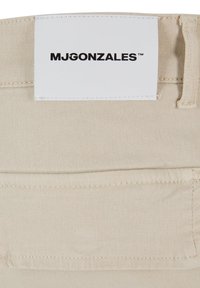 Beige színű pamut anyag, amelyen látható a textúra, valamint egy fehér címke található, rajta fekete betűkkel "MJGONZALES" felirattal, amely a derékpántra van varrva.