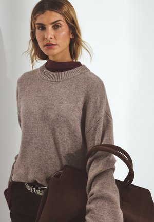 Woman wearing beige sweater and burgundy shirt, holding large brown leather handbag against plain white background.