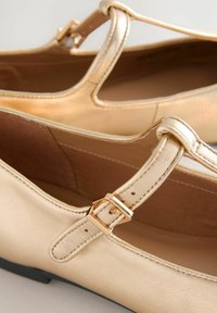 Zapatos estilo Mary Jane de cuero beige con correas de hebilla dorada y suelas planas negras, mostrados en detalle de primer plano.